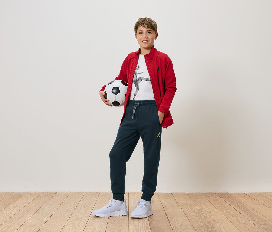 Un garçon tient un ballon de foot dans ses mains. Il porte une veste rouge, un pantalon et des baskets blanches.