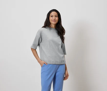 Femme aux cheveux foncés portant un haut gris et un pantalon bleu, la main dans la poche.