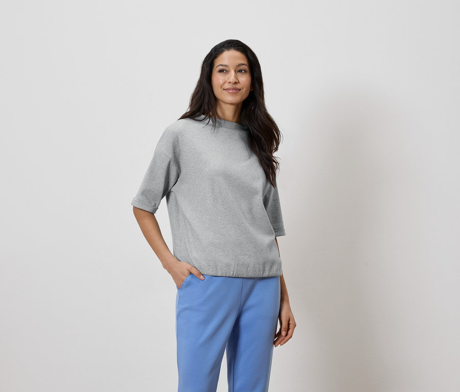 Femme aux cheveux foncés portant un haut gris et un pantalon bleu, la main dans la poche.