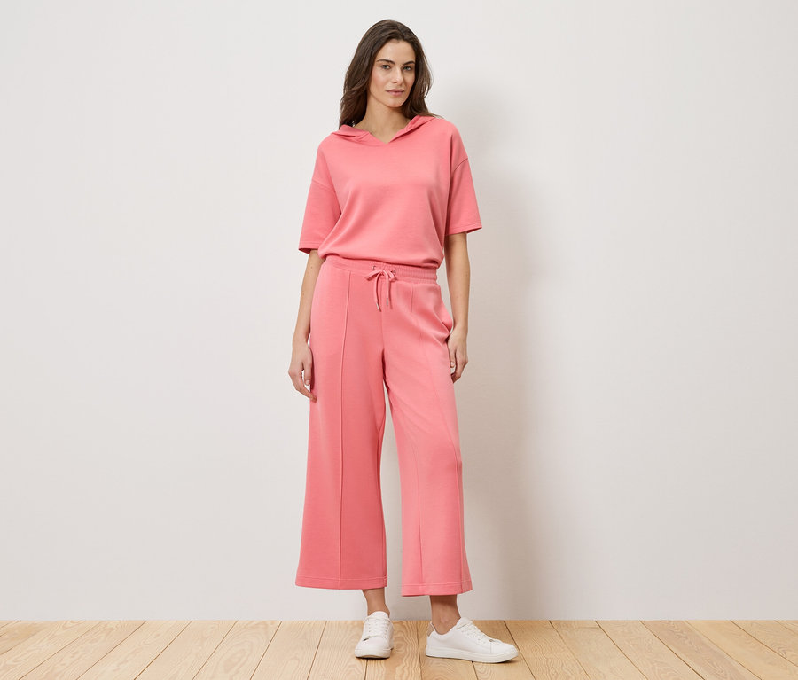 Une femme se tient devant un mur et porte un ensemble deux pièces corail composé d'un sweat à capuche à manches courtes et d'un pantalon culotte, ainsi que des baskets blanches.