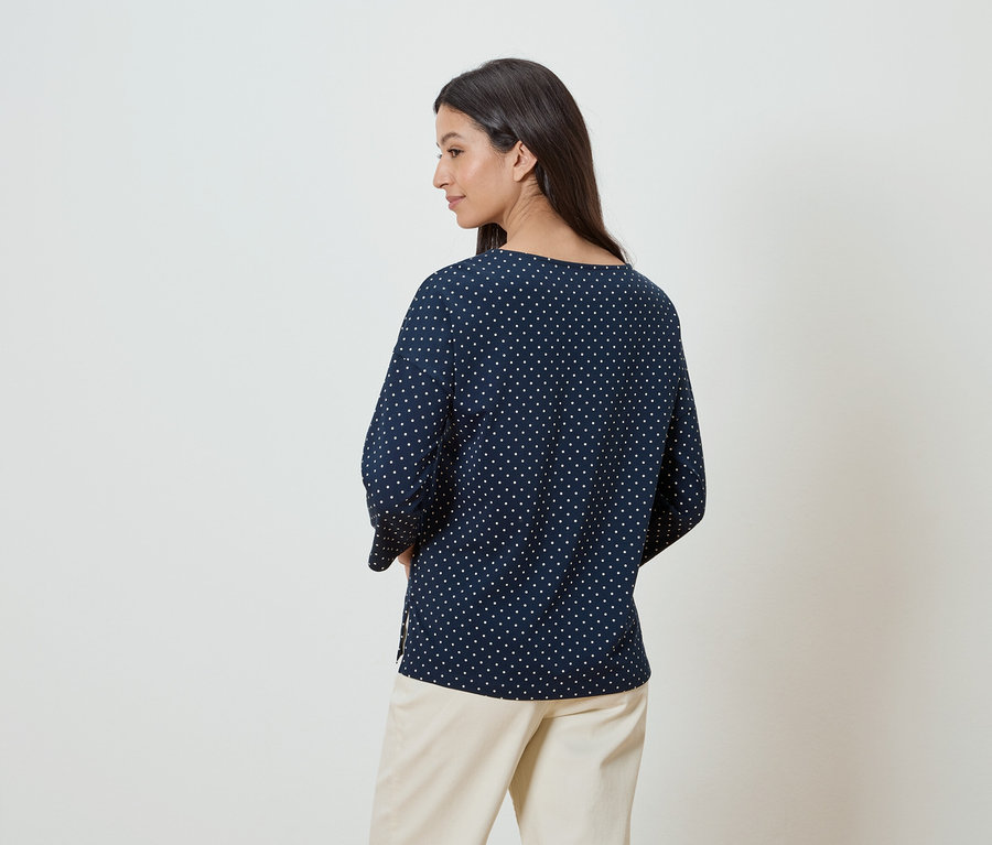 Une femme aux cheveux foncés porte un chemisier bleu marine à pois blancs et un pantalon beige.