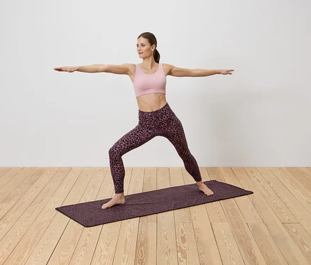 Une femme en tenue de sport fait du yoga sur un tapis à motifs sur un plancher en bois devant un mur blanc.