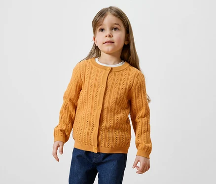 Une fillette aux longs cheveux châtain clair porte un cardigan doré et un jean.