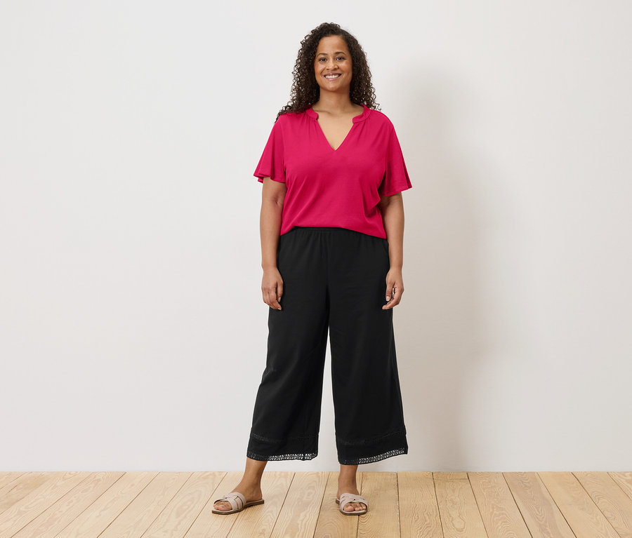 Une femme aux cheveux bouclés porte un t-shirt rose, une culotte noire avec des détails en dentelle et des sandales devant un mur blanc.