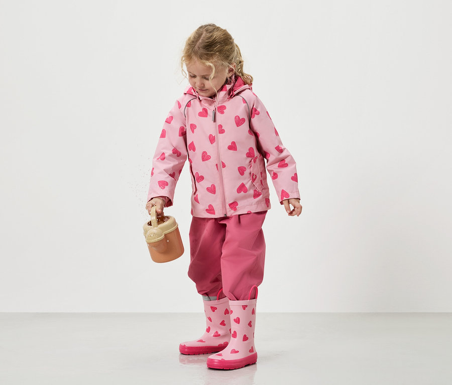 Une petite fille verse du sable d'un seau de jouet, vêtue d'une veste rose à motifs de cœur, d'un pantalon assorti et de bottes en caoutchouc.