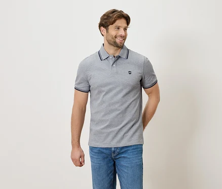 Homme portant un polo gris et un jean, debout devant un fond neutre.