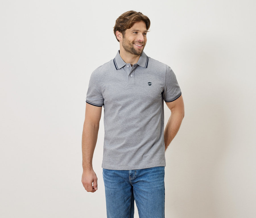 Homme portant un polo gris et un jean, debout devant un fond neutre.