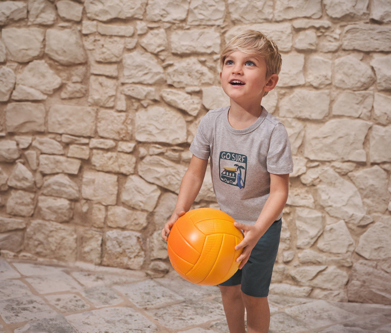 Un garçon tient un ballon orange et porte un pyjama court pour enfant SCHIESSER devant un mur de pierre.