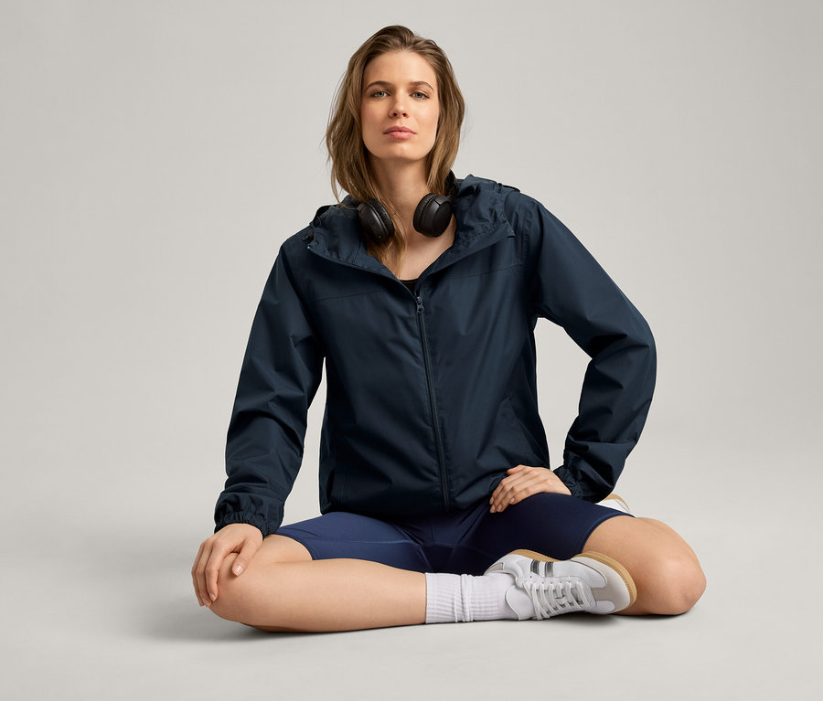 Femme assise en tailleur portant une veste de pluie, un casque audio, un short et des baskets.