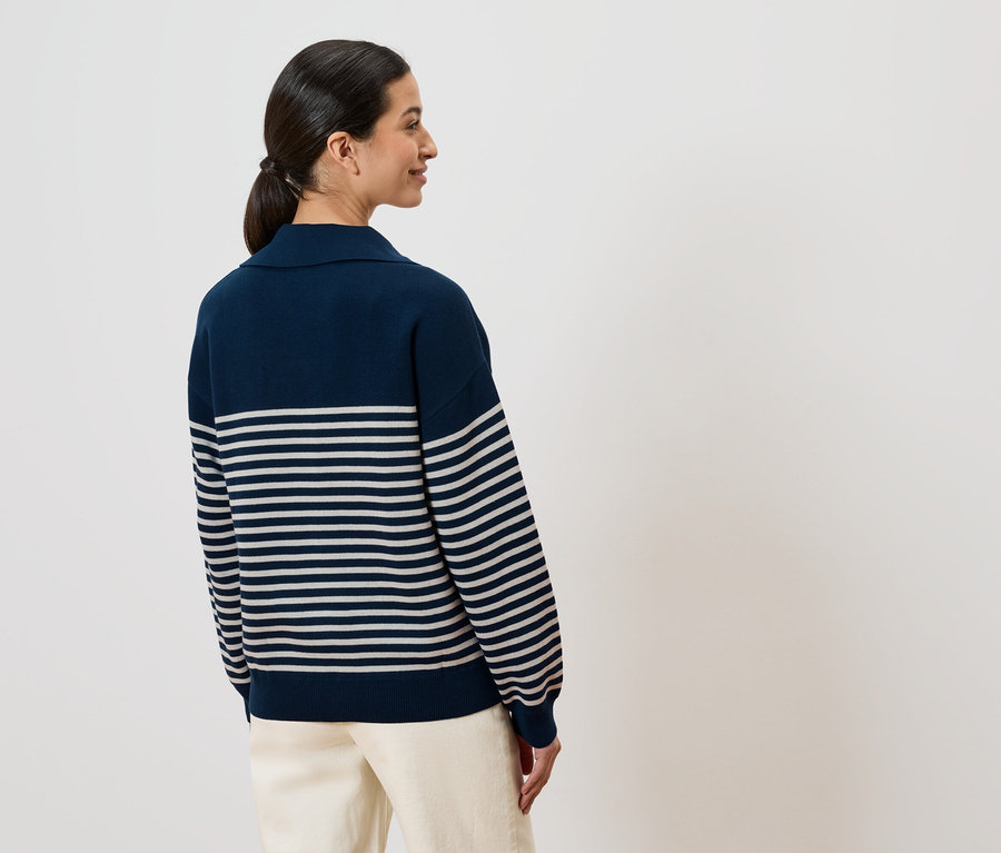 Femme de dos, queue de cheval, portant un pull rayé bleu marine et un pantalon clair sur fond blanc.