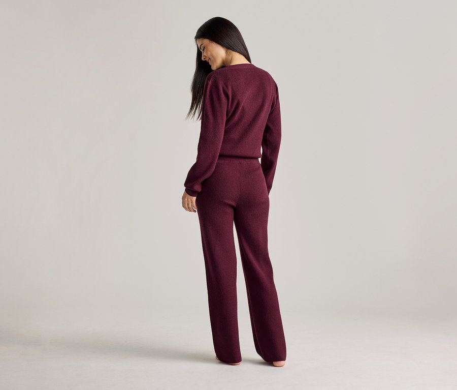 Une femme aux longs cheveux foncés se tient dos à la caméra, portant un haut et un pantalon en tricot côtelé bordeaux.