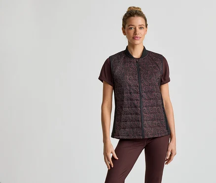 Une femme portant un chignon porte un t-shirt marron, un pantalon et un gilet matelassé zippé à motif léopard sur un fond clair.