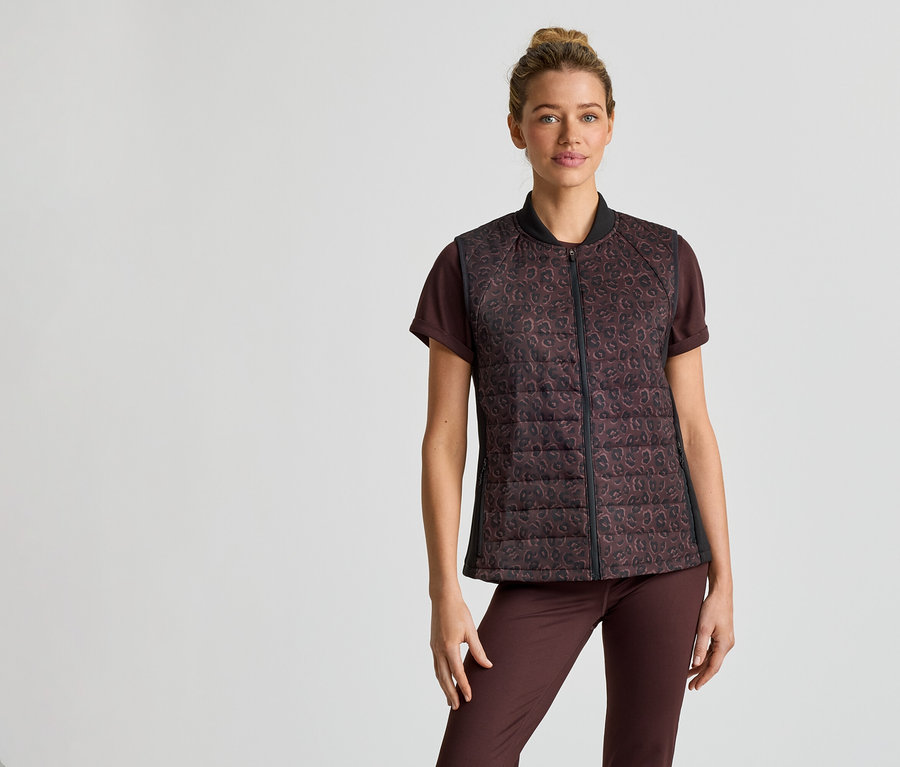 Une femme portant un chignon porte un t-shirt marron, un pantalon et un gilet matelassé zippé à motif léopard sur un fond clair.