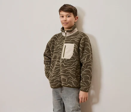 Un jeune garçon porte une veste polaire vert olive avec des motifs ondulés clairs et un jean clair, debout devant un mur clair. La veste a une fermeture éclair intégrale et une poche poitrine claire avec une fermeture éclair.