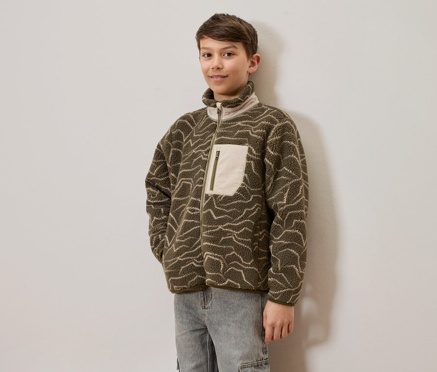 Un jeune garçon porte une veste polaire vert olive avec des motifs ondulés clairs et un jean clair, debout devant un mur clair. La veste a une fermeture éclair intégrale et une poche poitrine claire avec une fermeture éclair.