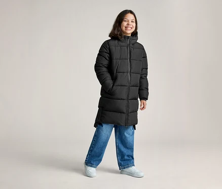 Une jeune personne souriante portant un long manteau matelassé noir, un jean bleu et des baskets gris clair, les mains dans les poches.