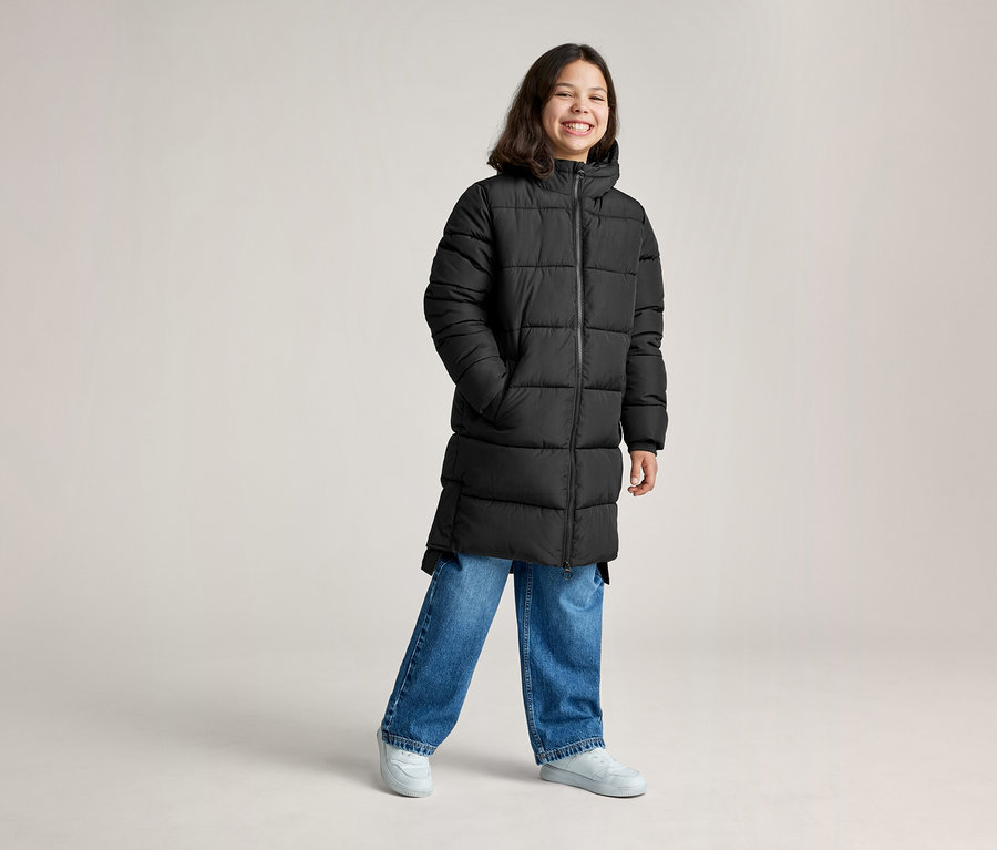 Une jeune personne souriante portant un long manteau matelassé noir, un jean bleu et des baskets gris clair, les mains dans les poches.