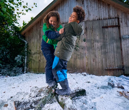 Deux enfants en vestes d'hiver et pantalons de neige rient en se tenant l'un à l'autre, en équilibre sur une bûche enneigée. Un abri en bois se trouve à l'arrière-plan.