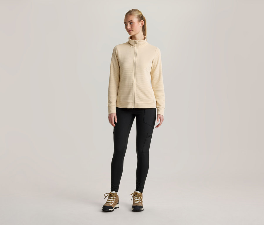 Une jeune femme est debout de face, vêtue d'une veste zippée jaune clair, d'un legging noir avec des poches latérales et de bottes de randonnée marron avec des lacets blancs. Ses cheveux blonds sont attachés en queue de cheval.