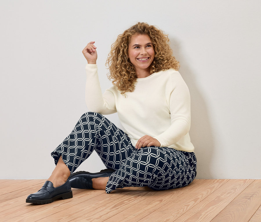 Femme aux cheveux bouclés assise sur un plancher en bois, portant un pull crème et un pantalon bleu à motifs géométriques.