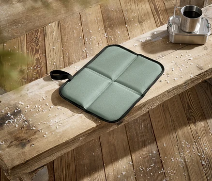 Un coussin de siège pliable vert clair à quatre sections repose sur un banc en bois rustique. À sa droite, une boîte à lunch en métal argenté avec une tasse en métal remplie de café. Des flocons blancs ressemblant à de la neige sont éparpillés sur le banc et le plancher en bois.