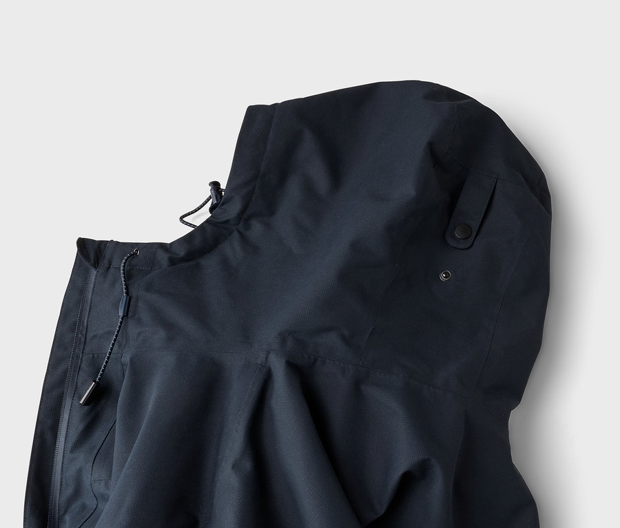 Gros plan d'une veste à capuche bleu foncé avec un cordon de serrage et un stoppeur près du col, et deux boutons-pression noirs sur le côté de la capuche.