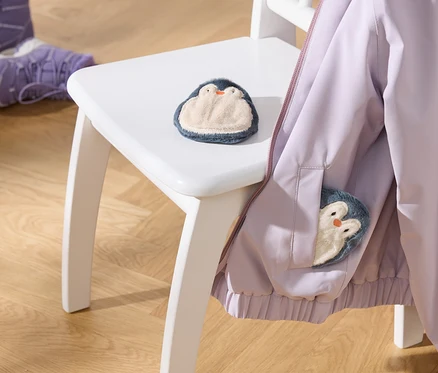 Une veste violet clair est suspendue à une chaise blanche, avec un jouet pingouin en peluche sur l'assise et un autre dans la poche de la veste. Deux bottes d'enfant violettes sont posées sur le parquet.