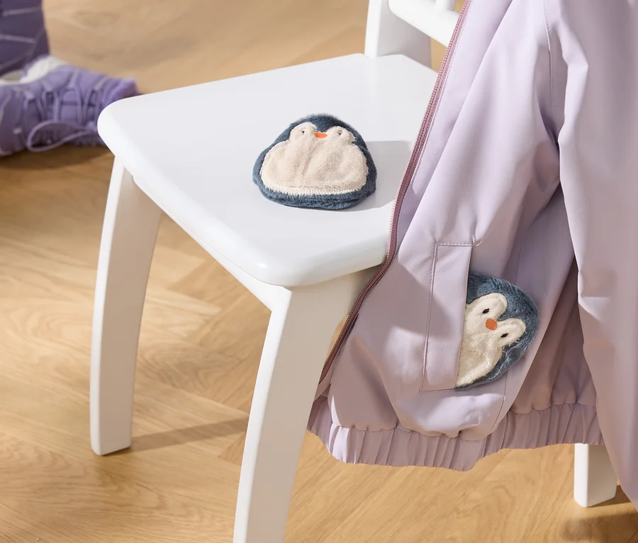 Une veste violet clair est suspendue à une chaise blanche, avec un jouet pingouin en peluche sur l'assise et un autre dans la poche de la veste. Deux bottes d'enfant violettes sont posées sur le parquet.