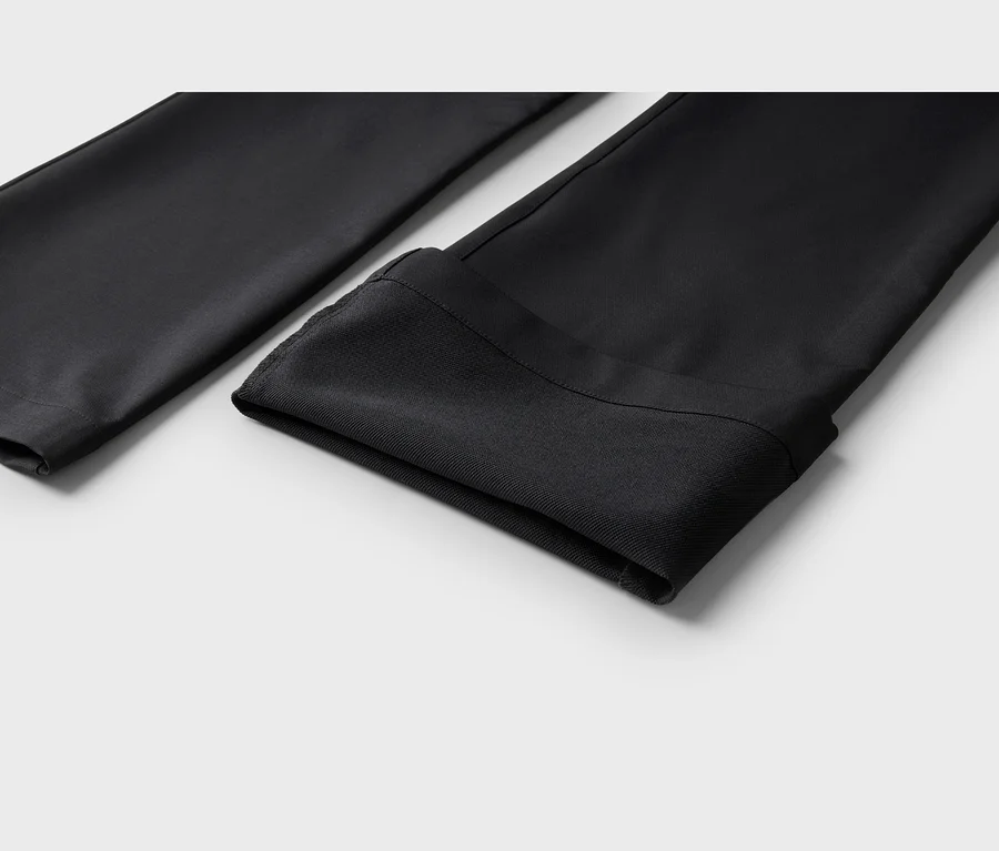 Deux pièces de tissu noir, probablement des jambes de pantalon ou des manches, reposent sur une surface gris clair. La pièce de droite a un revers plié, montrant une texture légèrement différente à l'intérieur.