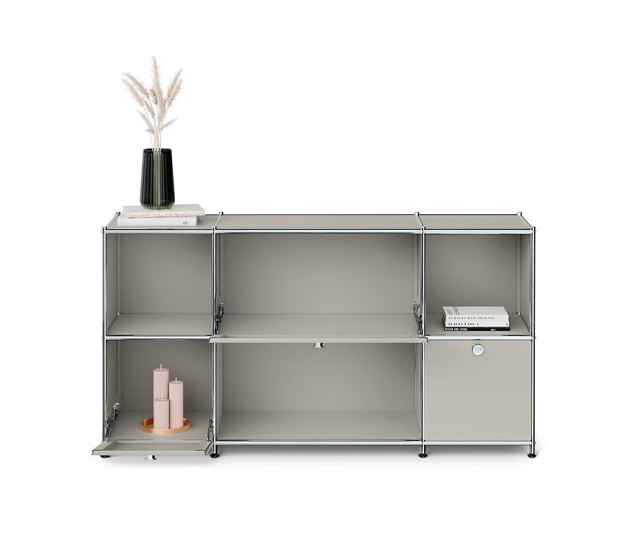 Unité d'étagères modulaires grise avec plusieurs compartiments. En haut à gauche, un vase avec de l'herbe de la pampa et des livres, en bas à gauche, trois bougies roses sur un plateau. À droite, un tiroir fermé.