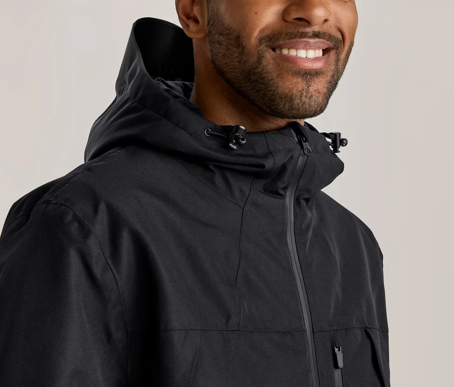 Homme souriant avec une barbe, portant une veste noire à capuche avec une fermeture éclair et des cordons de serrage.