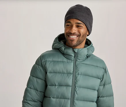 Un homme souriant portant un bonnet gris foncé et une doudoune vert clair à capuche regarde vers la droite.