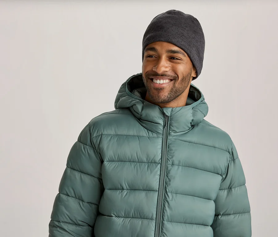 Un homme souriant portant un bonnet gris foncé et une doudoune vert clair à capuche regarde vers la droite.