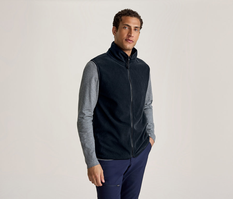 Homme portant un gilet polaire noir sur un t-shirt gris à manches longues et un pantalon bleu marine, debout une main dans la poche.