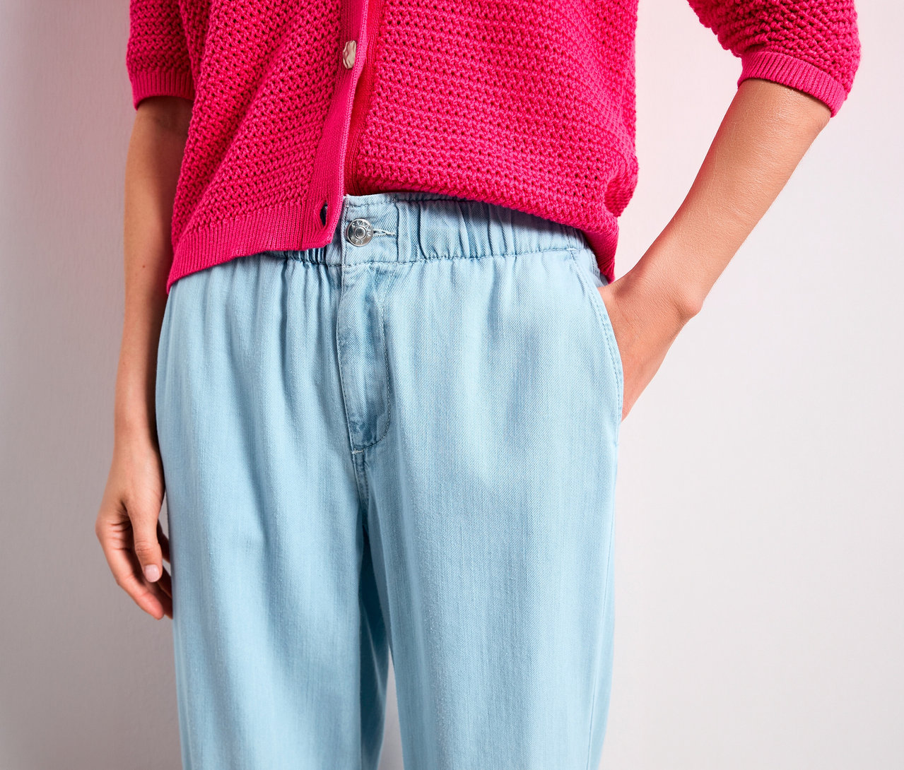 Une personne porte un cardigan en maille rose vif et un jean Street One « Emee » bleu clair avec une taille élastique. Une main est dans la poche du jean.