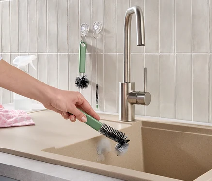 Une main nettoie un évier de cuisine beige avec une brosse verte. Un robinet en acier inoxydable, des carreaux muraux clairs, une serviette rose et d'autres brosses de nettoyage sont visibles.