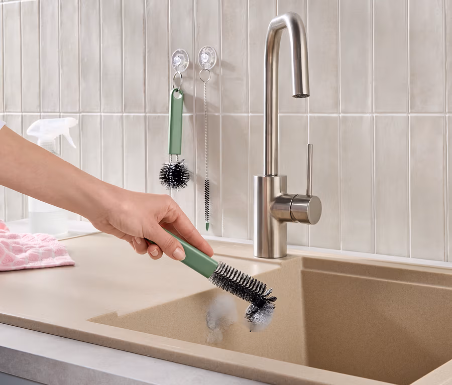 Une main nettoie un évier de cuisine beige avec une brosse verte. Un robinet en acier inoxydable, des carreaux muraux clairs, une serviette rose et d'autres brosses de nettoyage sont visibles.