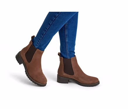 Personne portant un jean bleu et des bottines Chelsea en daim marron avec des empiècements élastiques latéraux et une semelle épaisse, sur fond blanc.