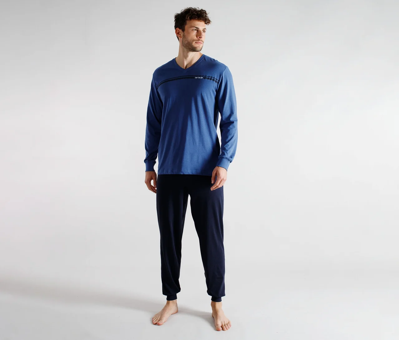 Un homme portant un t-shirt bleu à manches longues et col en V avec un détail de rayure horizontale et un pantalon de survêtement bleu foncé, debout pieds nus. Il regarde vers la droite.