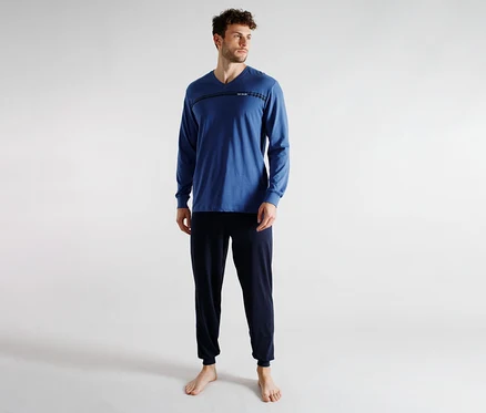 Un homme portant un t-shirt bleu à manches longues et col en V avec un détail de rayure horizontale et un pantalon de survêtement bleu foncé, debout pieds nus. Il regarde vers la droite.