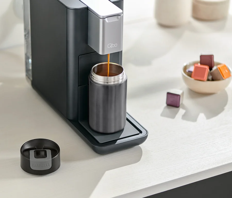Machine à café remplissant un gobelet gris Kambukka. Le couvercle du gobelet est à côté de la machine à café. Un bol de capsules est à côté.