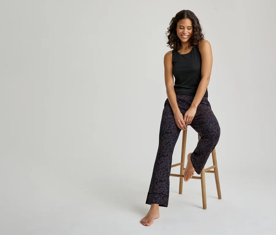 Une femme souriante aux cheveux bouclés foncés est assise sur un tabouret en bois. Elle porte un débardeur noir et un pantalon de pyjama foncé à motifs. Ses pieds sont nus.