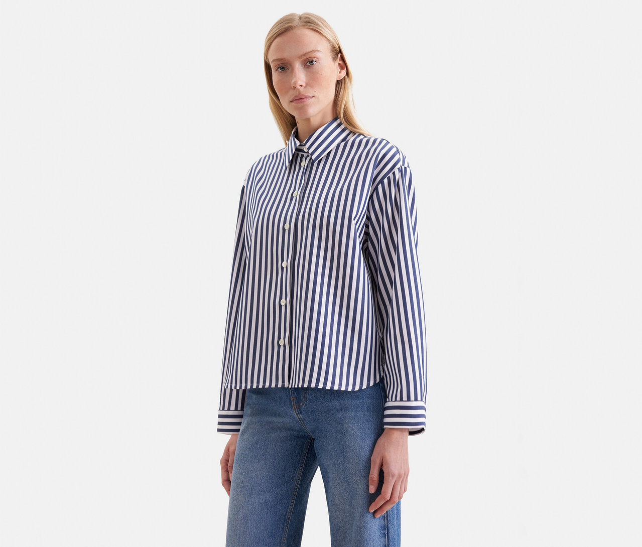 Femme blonde portant une chemise à manches longues rayée bleu marine et blanc et un jean bleu, regardant devant elle.