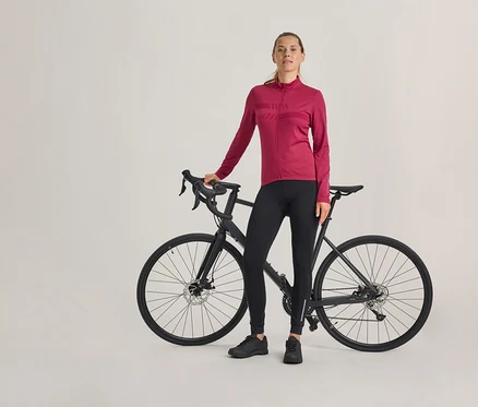 Femme debout à côté d'un vélo de route noir, portant un maillot de cyclisme fuchsia à manches longues, un pantalon de cyclisme noir et des chaussures noires.