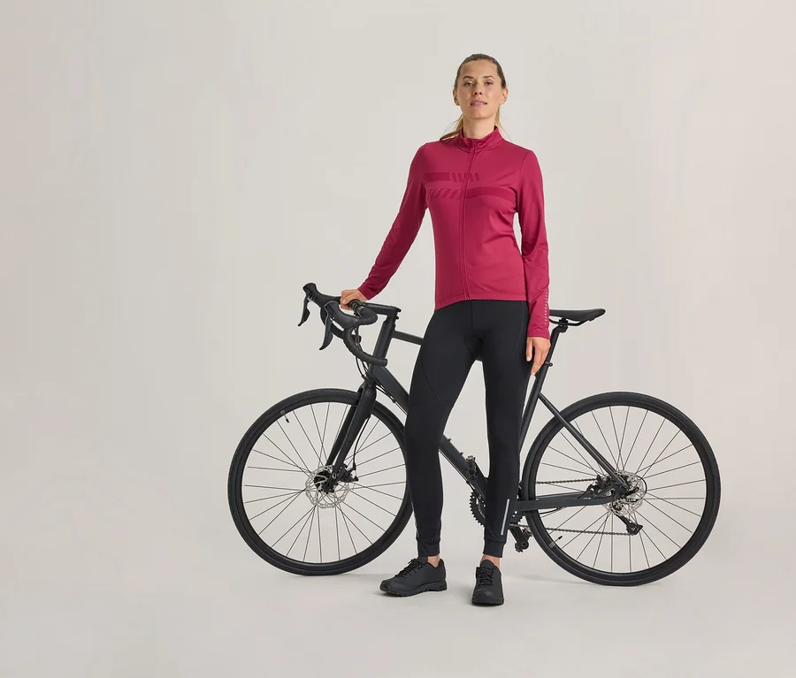 Femme debout à côté d'un vélo de route noir, portant un maillot de cyclisme fuchsia à manches longues, un pantalon de cyclisme noir et des chaussures noires.