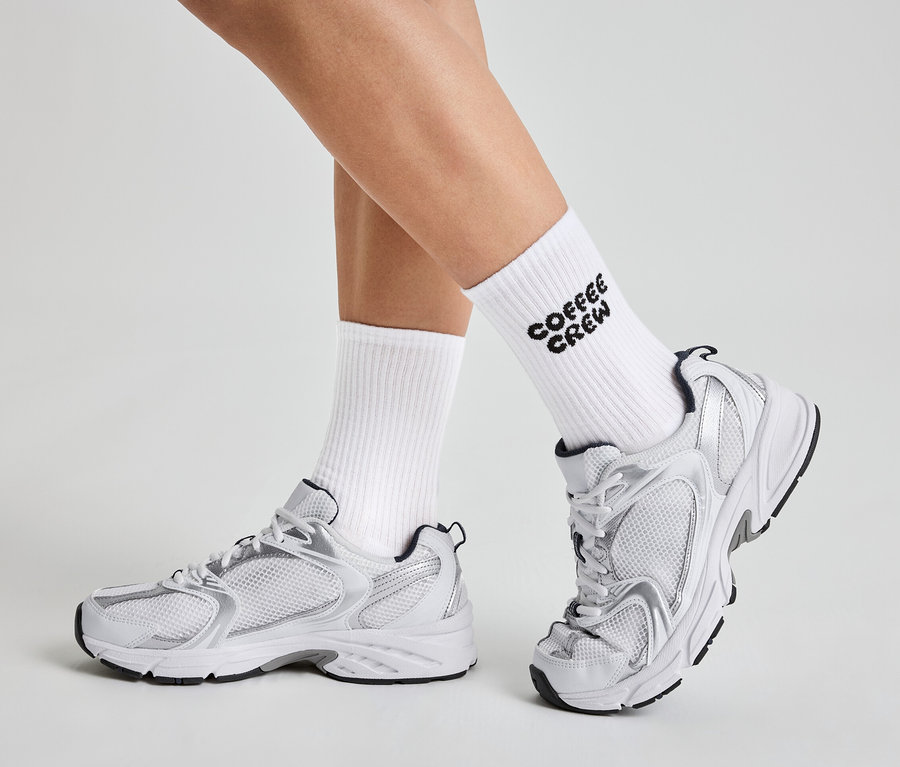 Jambes d'une personne portant des chaussettes blanches avec l'inscription "Coffee Crew" et des baskets blanches et argentées.