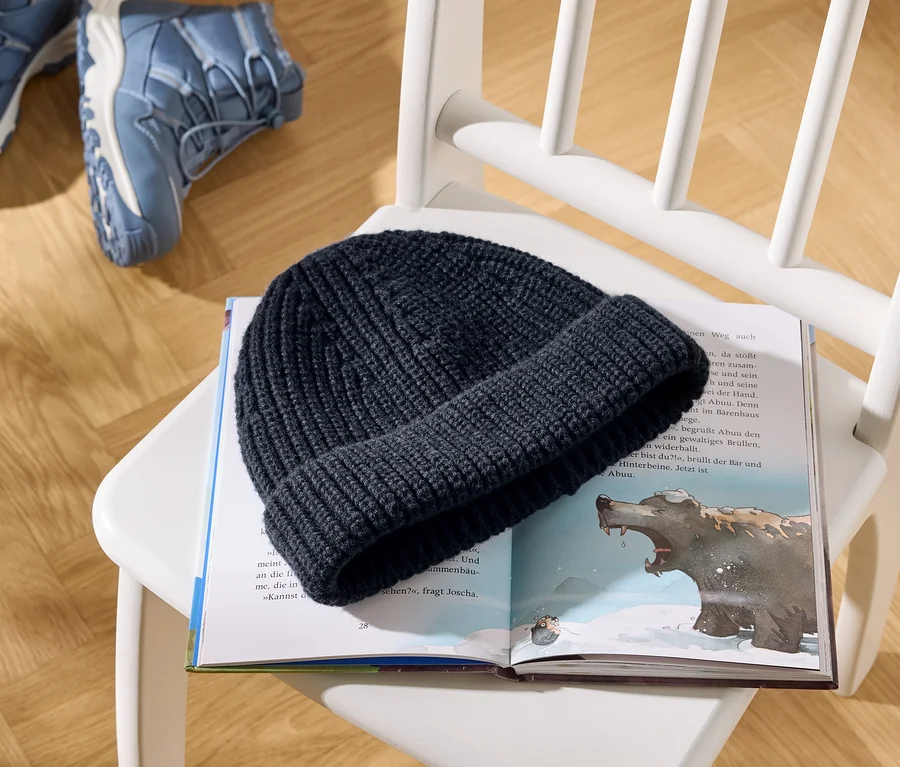 Un bonnet tricoté bleu foncé repose sur un livre pour enfants ouvert, illustrant un ours et du texte. Le livre est posé sur une chaise en bois blanche, avec deux chaussures de randonnée bleues visibles sur le parquet en arrière-plan.
