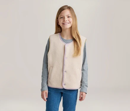 Jeune fille souriante aux longs cheveux blonds, portant un gilet polaire crème avec des bordures violettes sur un t-shirt gris à manches longues et un jean bleu.