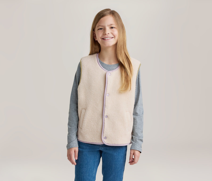 Jeune fille souriante aux longs cheveux blonds, portant un gilet polaire crème avec des bordures violettes sur un t-shirt gris à manches longues et un jean bleu.