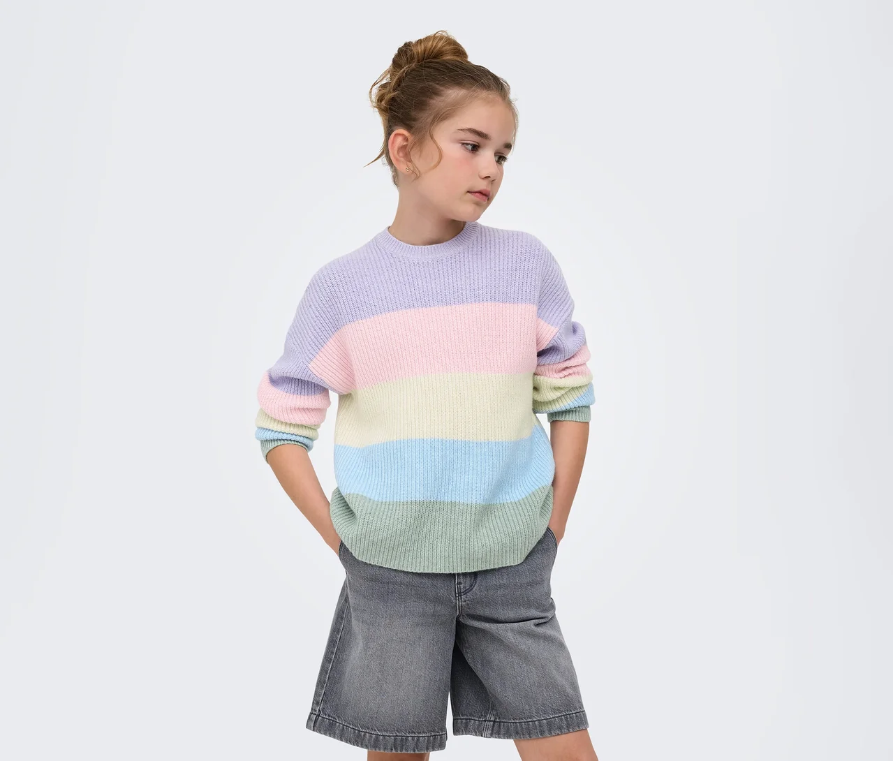 Jeune fille aux cheveux en chignon, portant un pull en maille rayé ONLY GIRLS Sandy lavande, rose, jaune, bleu clair et vert clair, et un short en jean gris, mains dans les poches, regardant sur le côté devant un fond blanc.